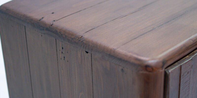 A close-up view of the top corner of a wooden dresser, showcasing its rich brown finish and natural wood grain texture. The surface has visible seams, knots, and small imperfections, adding to its rustic charm. The dresser's rounded edges and smooth finish highlight its craftsmanship. The background is a plain white surface.